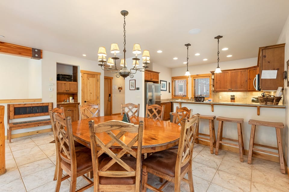 A kitchen and dining area with a wooden dining table and chairs, a kitchen island with bar stools, wooden cabinets, hanging pendant lights, and modern stainless steel appliances.