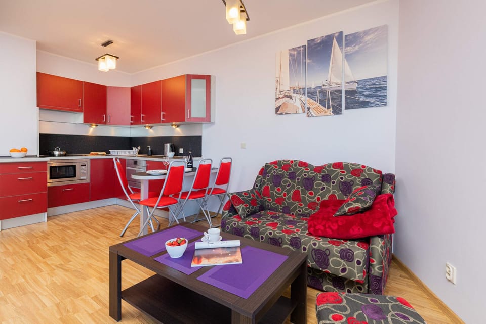 Open-plan living and dining area with red kitchen accents, wooden floors, and modern lighting.
