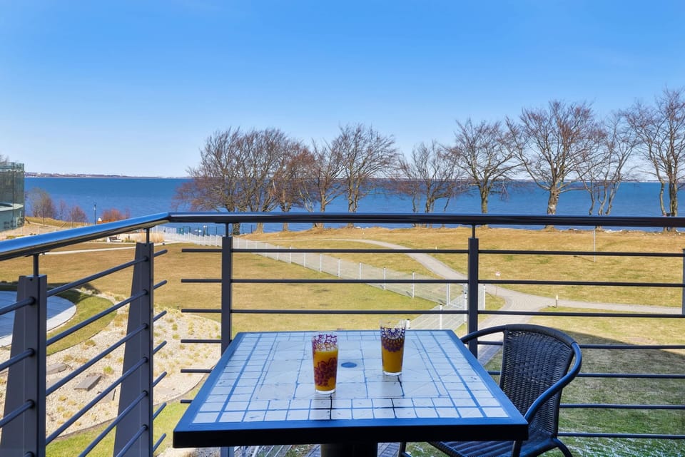 Balcony with a table and chairs offering a peaceful view of the waterfront and green landscape.
