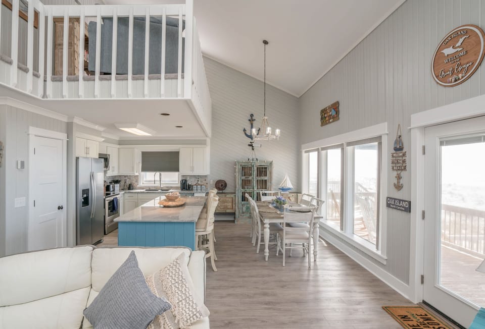 Overhead view of a bright beach house living room with white sectional seating, striped rug, blue side tables, TV console, ceiling fan, and glass doors opening to a covered porch.