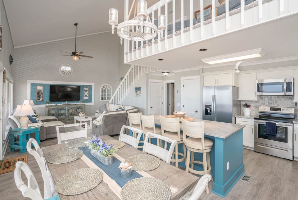 Bright dining area with table for six, distressed beach-style cabinet, bar seating, chandelier, and large windows overlooking the covered deck and Atlantic view.