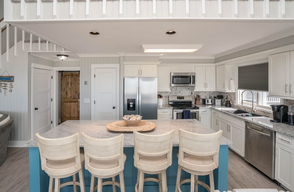 Open kitchen with a blue island, four bar stools, white cabinets, stainless appliances, dining table, distressed beach-style cabinet, loft railing above, and light wood-look floors.
