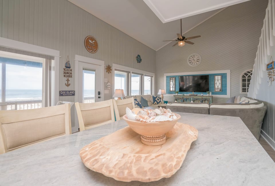 Bright dining area with table for six, distressed beach-style cabinet, bar seating, chandelier, and large windows overlooking the covered deck and Atlantic view.