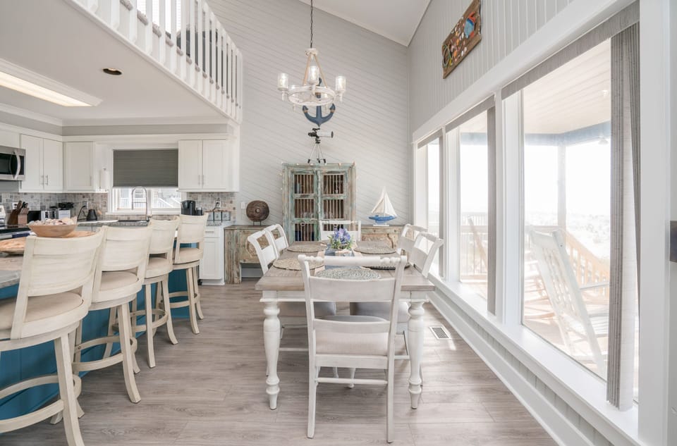 Bright dining area with table for six, distressed beach-style cabinet, bar seating, chandelier, and large windows overlooking the covered deck and Atlantic view.