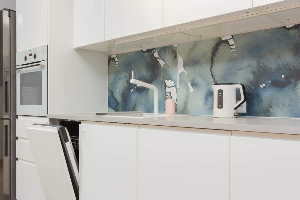 A modern kitchenette with sleek white cabinets and a minimalist design. The backsplash features a decorative glass panel with artistic elements.