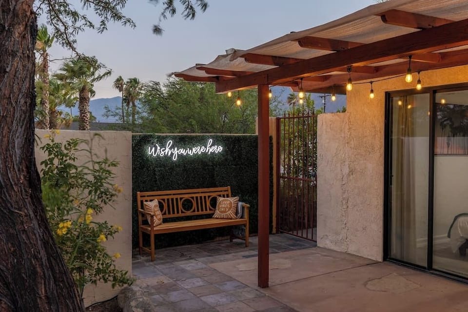 Twilight front courtyard with wood bench, string lights, pergola, and a neon 'Wish You Were Here' sign against mountain views