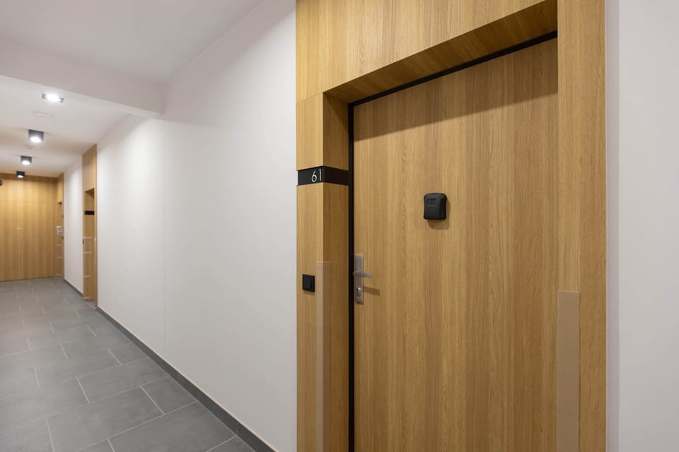 A hallway in the building with light wooden doors and tiled flooring, leading to the apartment entrance.







