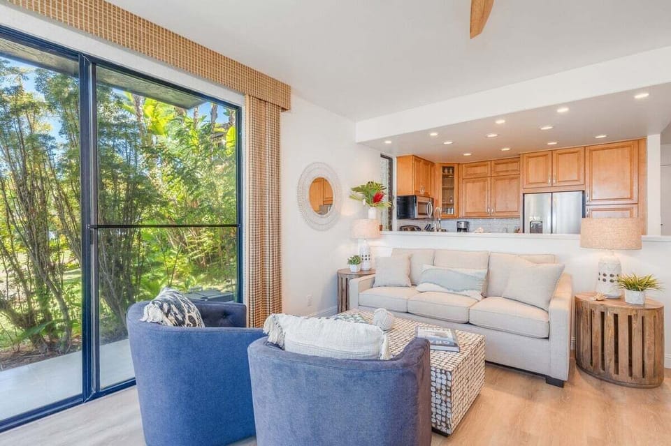 Cozy living room with light sofas, wicker accents, and blue armchairs opening to a kitchen with warm wood cabinetry