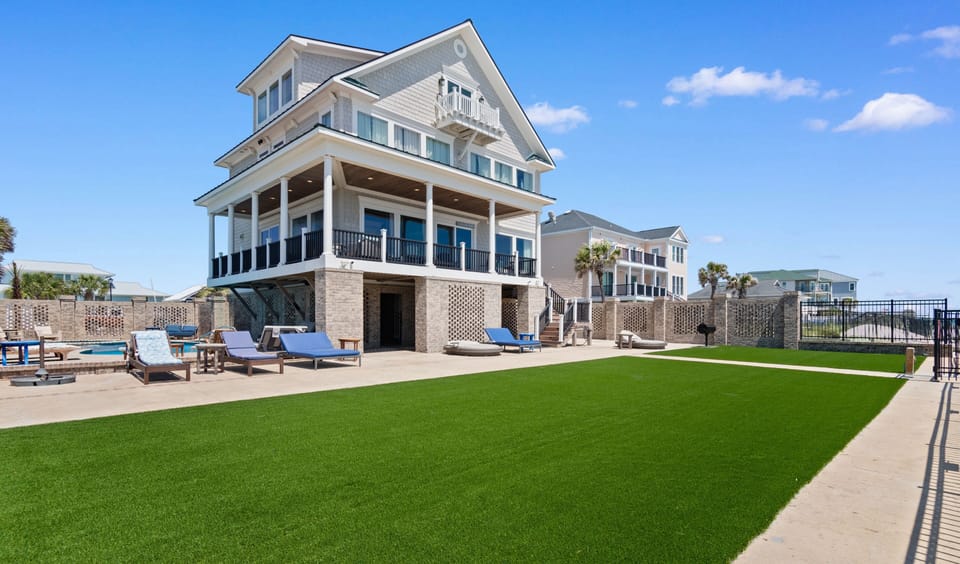 Modern coastal home featuring multiple levels with balconies, pool area, and pristine artificial lawn under clear blue skies.
