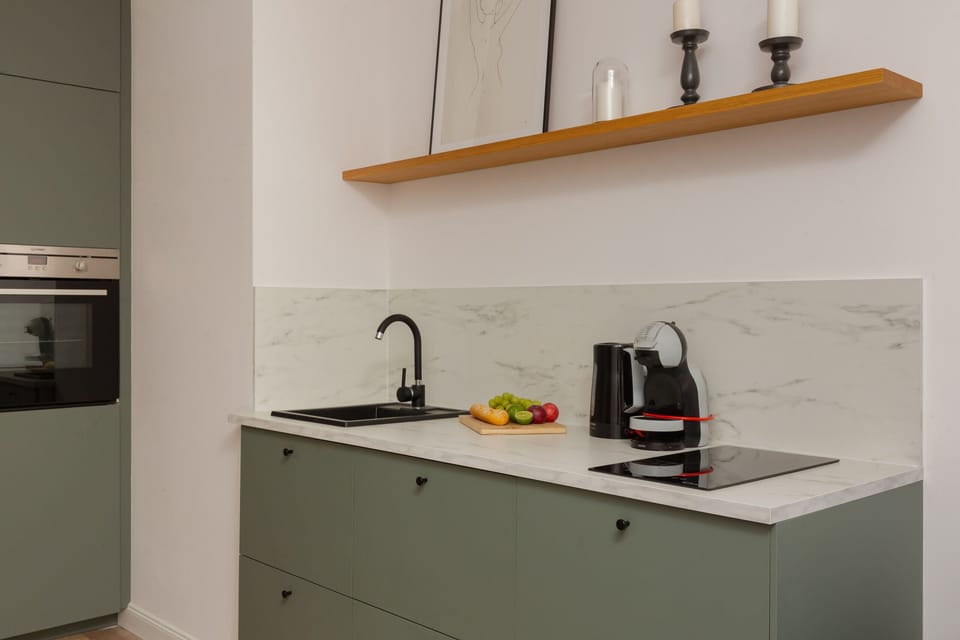 A compact kitchenette with green cabinets, a countertop, a coffee maker, and a minimalist wooden shelf.