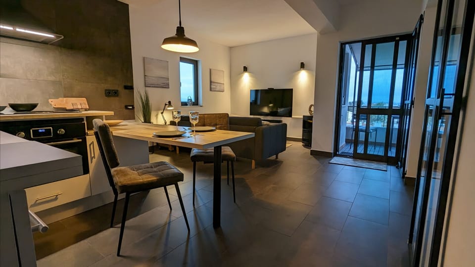 An open-plan, modern living area with an integrated kitchen, dining area, and comfortable seating area. The stylish furnishings in neutral tones create a pleasant atmosphere. The room is equipped with a flat-screen TV, comfortable sofas, and decorative lighting. Floor-to-ceiling glass doors lead directly to the terrace with its view, flooding the room with natural light. Ideal for relaxing evenings, cooking together, or cozy get-togethers.