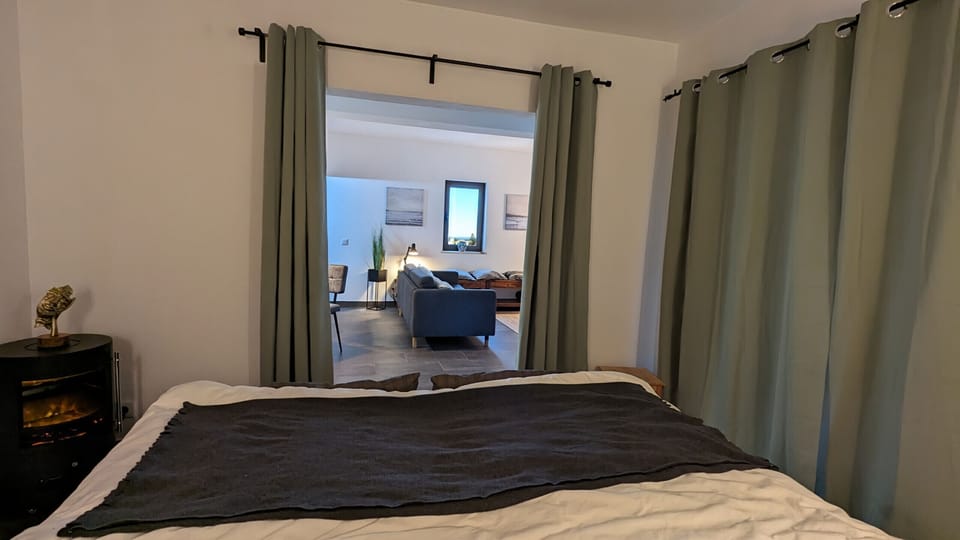The cozy bedroom features a large bed with a dark duvet and light-colored sheets. To the left of the bed is a nightstand with a golden sculpture and a wood-burning stove beneath it. A light olive-green room divider curtain separates the bedroom from the living area behind it. Through the opening, a sofa, a coffee table, and a window are visible. To the right of the bed, another curtain separates the room from another area.