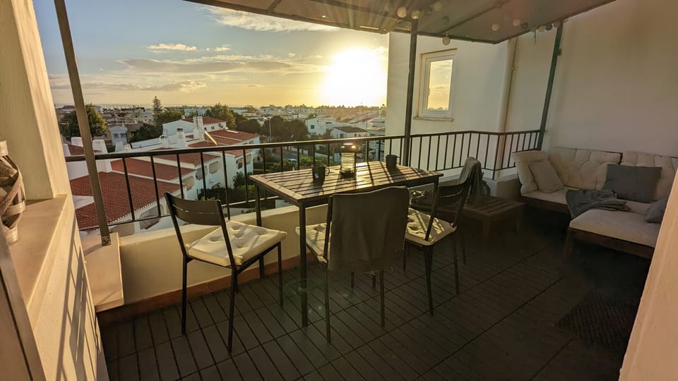 This spacious terrace offers breathtaking views of the surrounding countryside with its white houses and the distant sea, shimmering in the golden light of the sunset. In the foreground, a square dark wood dining table with four chairs is ready for al fresco meals. To the right, a comfortable seating area with a light-colored sofa and a side table invites relaxation. The floor is laid with dark wood tiles, and a pergola provides shelter from the sun or light rain. Several small lights on the pergola create a cozy atmosphere in the evenings.