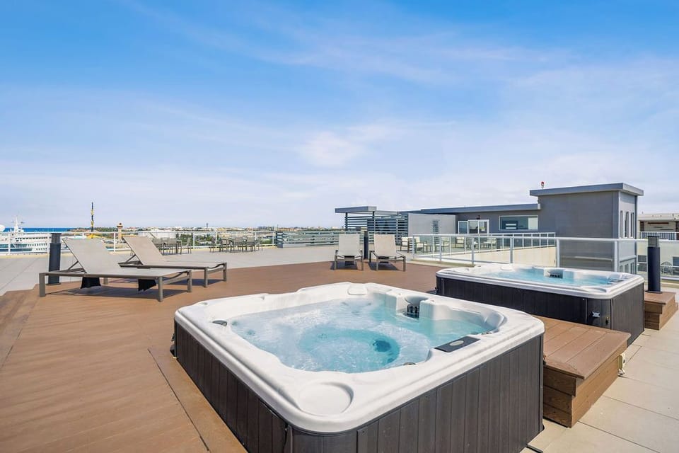 Modern apartment complex featuring a striking glass-walled pool in an inner courtyard. The pool area is surrounded by a well-maintained landscaped hedge, vibrant blue umbrellas, and loungers—ideal for relaxation under sunny skies.
