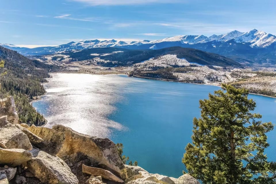 A large lake surrounded by snow-capped mountains and lush forests under a clear blue sky, seen from a rocky vantage point.
