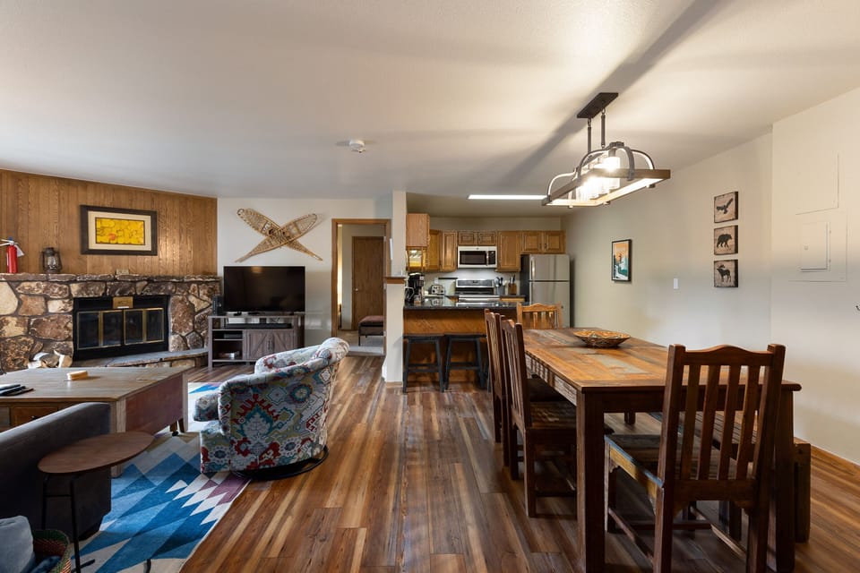 A cozy living and dining area with wood flooring, stone fireplace, TV, colorful rug, patterned armchair, wooden dining table with chairs, and a kitchen with stainless steel appliances in the background.
