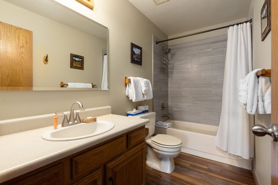 A bathroom with a sink and vanity, toilet, bathtub with shower, white curtain, and towel racks. The walls are beige, and the floor is wooden. A mirror and framed pictures are on the walls.