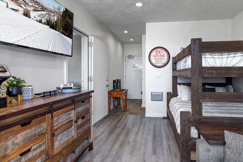 Your mountain-ready suite features rustic bunk beds and a wood dresser beneath a TV showing live slope views, blending cozy ski-lodge style with modern comfort.