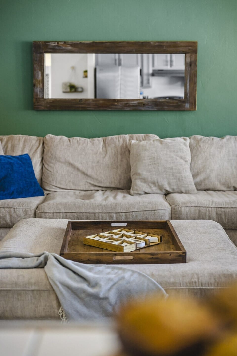 Living Room Lounge Detail: Unwind together on the plush sectional sofa for cozy movie nights and board game sessions, highlighted by soft accent pillows, a matching Ottoman footrest with a rustic wooden tray, and a framed mirror accenting the modern green