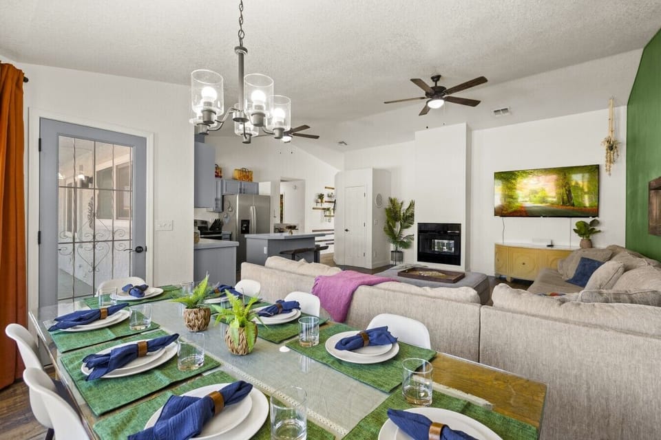 Open-Concept Living & Dining Area: Host and relax in this spacious open-concept living area, featuring a large dining table fully set for group meals, a plush sectional sofa, wall-mounted smart TV, modern fireplace, ceiling fan, and seamless view into the