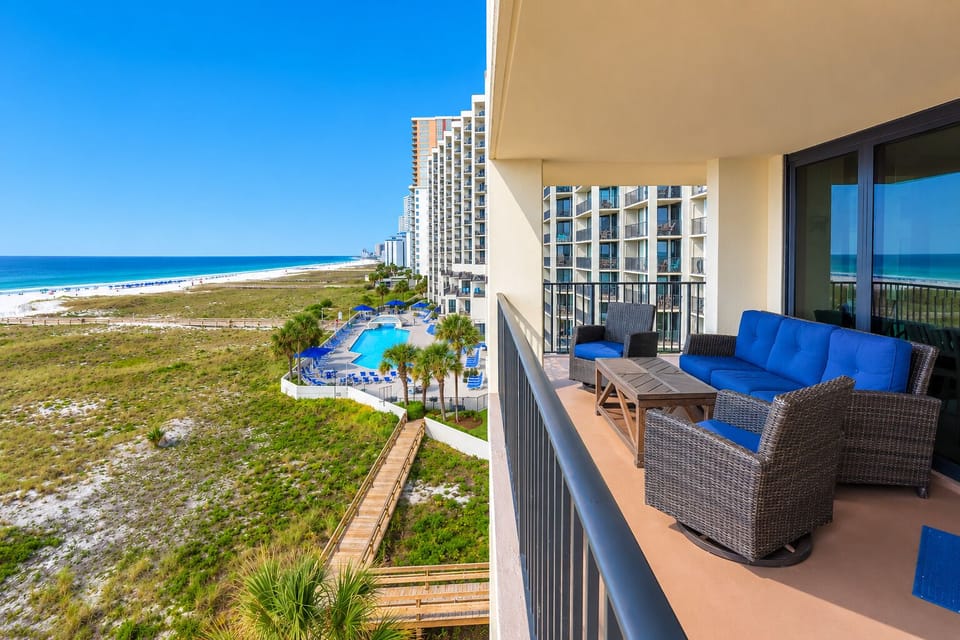 Spacious balcony features comfortable seating with vibrant blue cushions, offering a view of the beach and nearby pool area