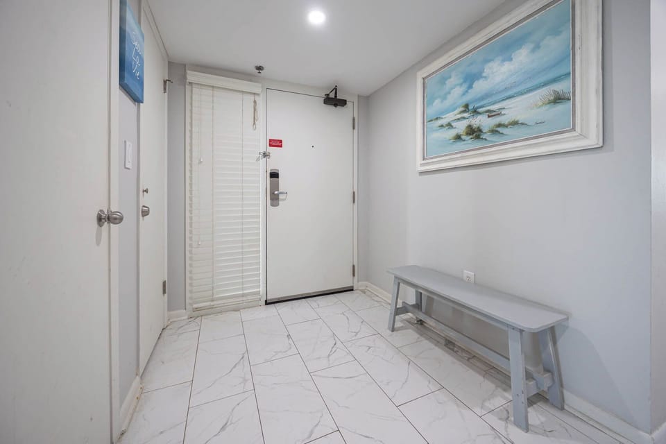 Welcoming entryway features light-colored tile flooring, a gray bench, and beach artwork, creating a relaxed atmosphere