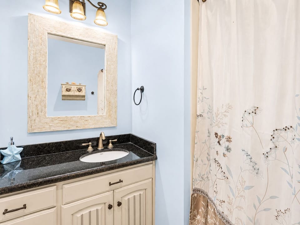 Guest bathroom with a granite vanity, cabinet storage, framed mirror, decorative lighting, and a shower-tub combination.