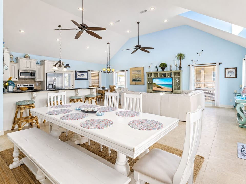 Open dining and living space with a large table, abundant seating, vaulted ceilings, ceiling fans, and bright coastal interiors.