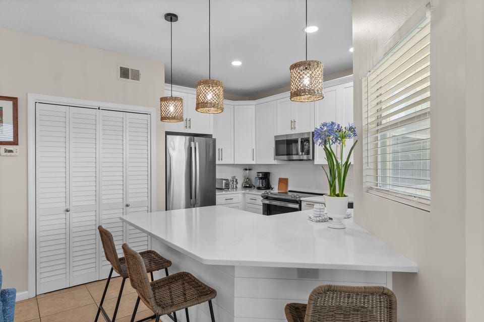 Your fully-equipped kitchen invites culinary adventures with gleaming white counters, modern appliances, and warm wicker pendant lights creating the perfect cooking atmosphere.