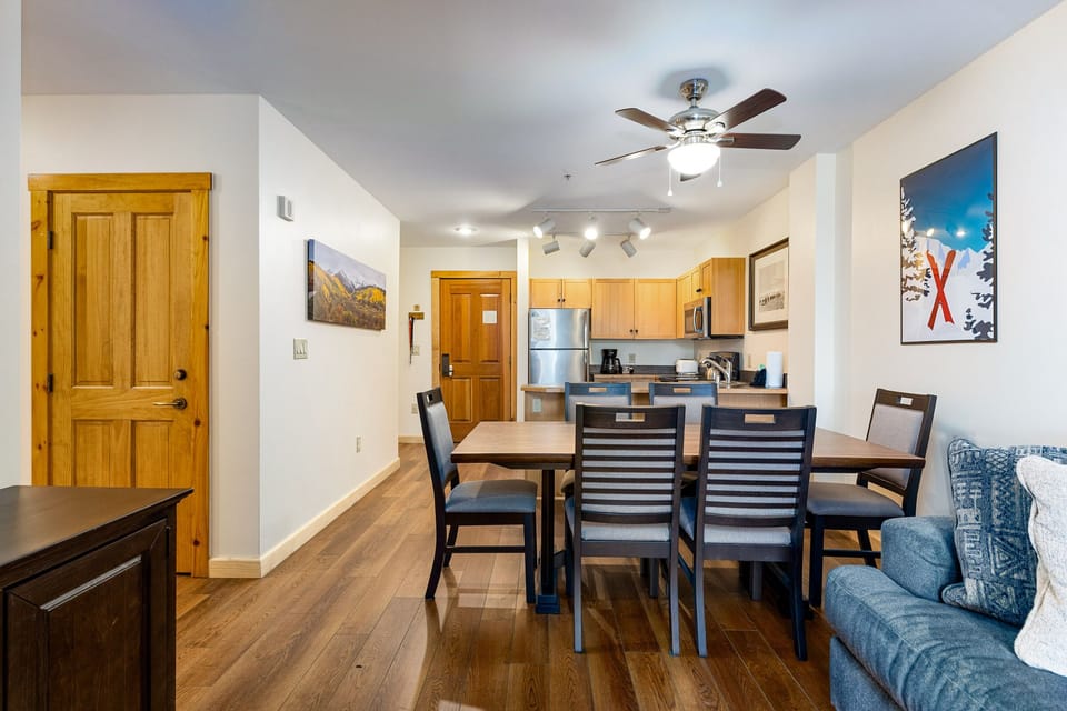 A modern kitchen and dining area with a wooden dining table, six chairs, stainless steel appliances, wooden flooring, and wall art. A ceiling fan is above the table, and a sofa is partially visible.