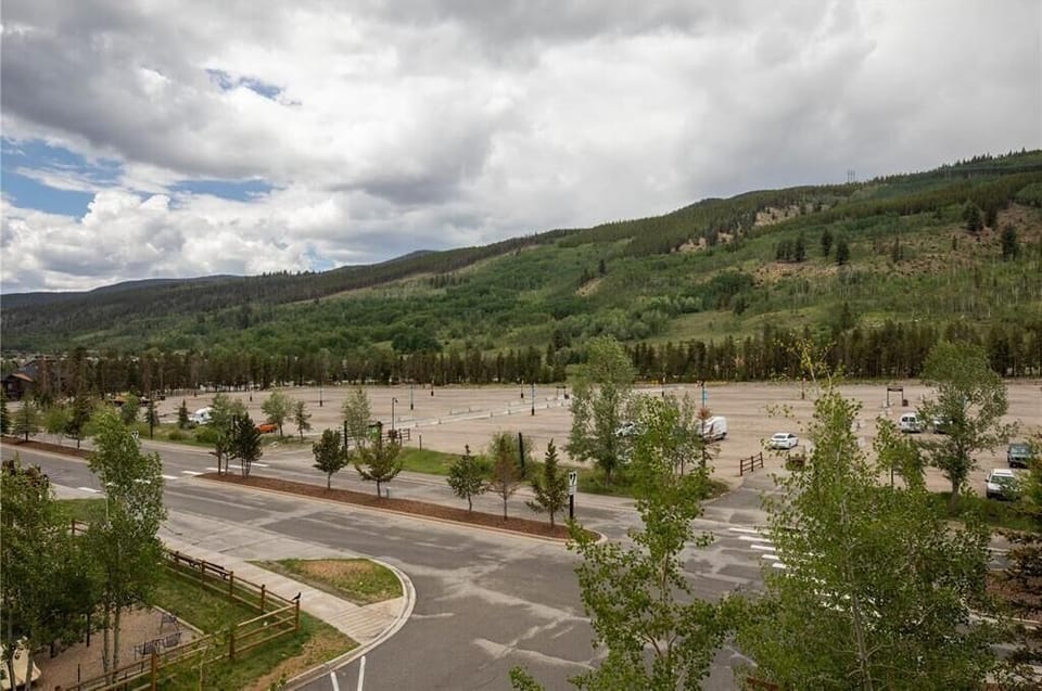 A large, mostly empty parking lot surrounded by trees and distant mountains under a cloudy sky. There are a few cars parked and trees lining the lot and the road in the foreground.