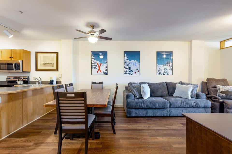 A living and dining area features a wooden dining table with chairs, a couch with cushions, wall art, a ceiling fan, and a kitchen with wooden cabinets and stainless steel appliances.