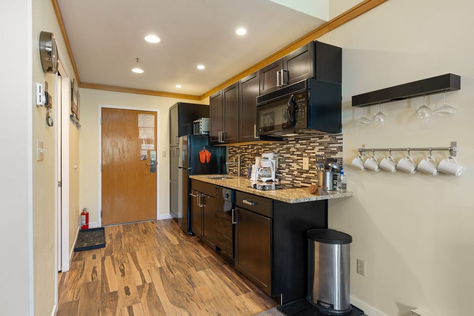 Modern kitchen with wood flooring, dark cabinetry, stainless steel appliances, granite countertops, and tile backsplash. Wall-mounted wine glass and mug racks are also visible.