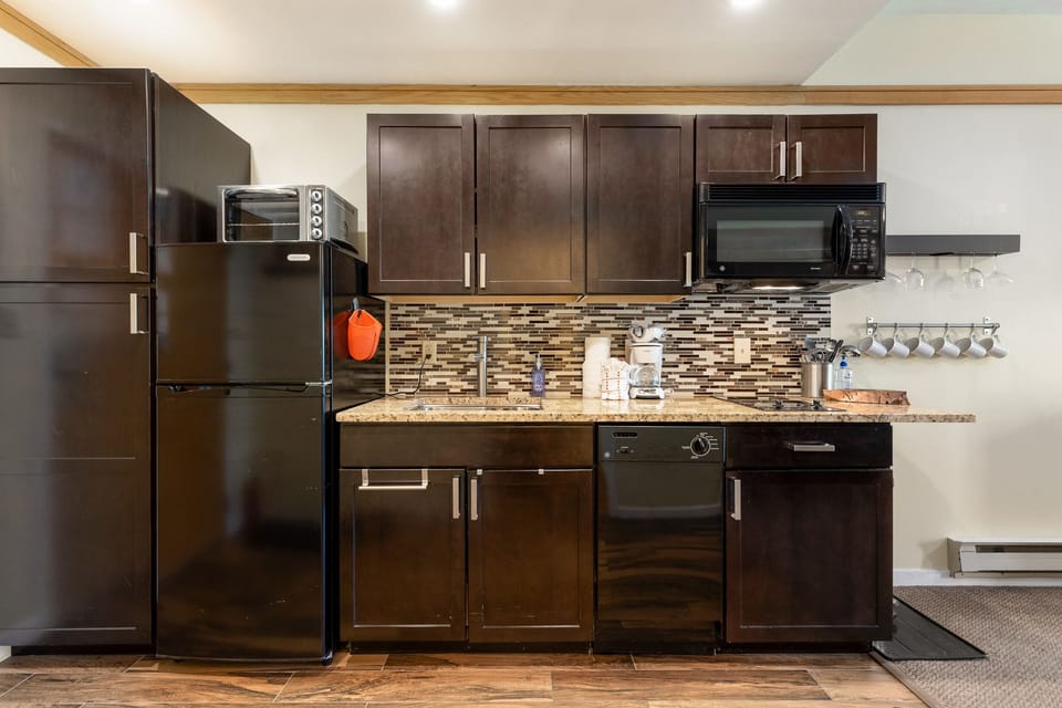 Modern kitchen with dark wood cabinets, black appliances including a refrigerator, microwave, and dishwasher, a granite countertop, and a tile backsplash. Cups and utensils are visible on the counter.
