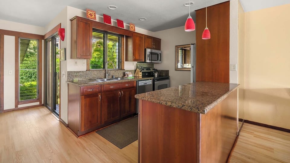 Step into this bright, fully-equipped kitchen with gleaming granite counters, rich wood cabinetry, and natural light streaming through glass doors that open to lush greenery beyond.