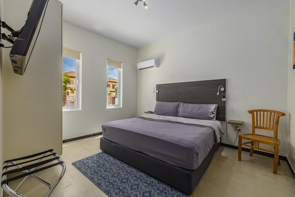 This bedroom features a king-size bed, a comfortable mattress, and an elegant bed headboard. There is an air conditioning unit, extra seating, and windows that allow natural light to get in.