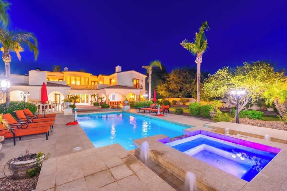 Expansive villa resort features illuminated pool and spa set amid lush gardens under dramatic twilight skies, with multiple outdoor lounging areas, tropical palms, and elegant Mediterranean-style architecture.