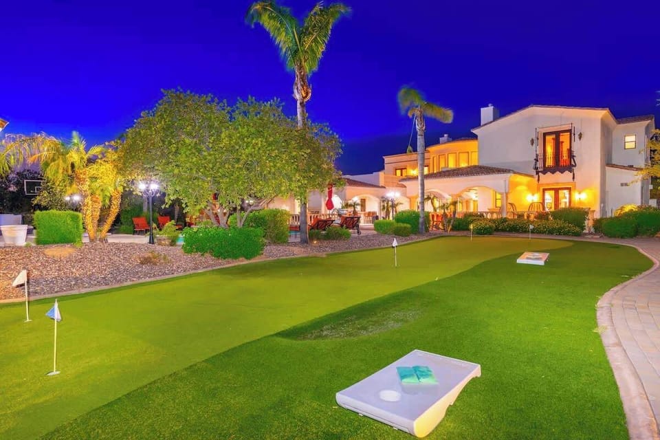 The property features a private putting green, perfect for friendly competition. Illuminated palm trees and manicured landscaping frame the white stucco villa, creating an inviting resort-style atmosphere at twilight.