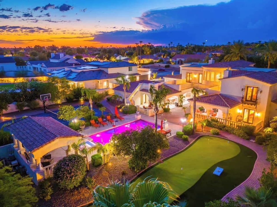 A stunning twilight aerial view captures this expansive luxury rental estate featuring a sparkling pool, private putting green, and elegant outdoor entertaining spaces set within a beautifully landscaped desert neighborhood.