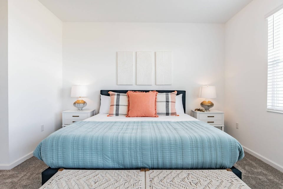 Experience comfort in this cozy bedroom with a plush king-sized bed dressed in soft blue and coral linens. Modern nightstands and lamps add warmth, while natural light from the large window enhances the tranquil ambiance.