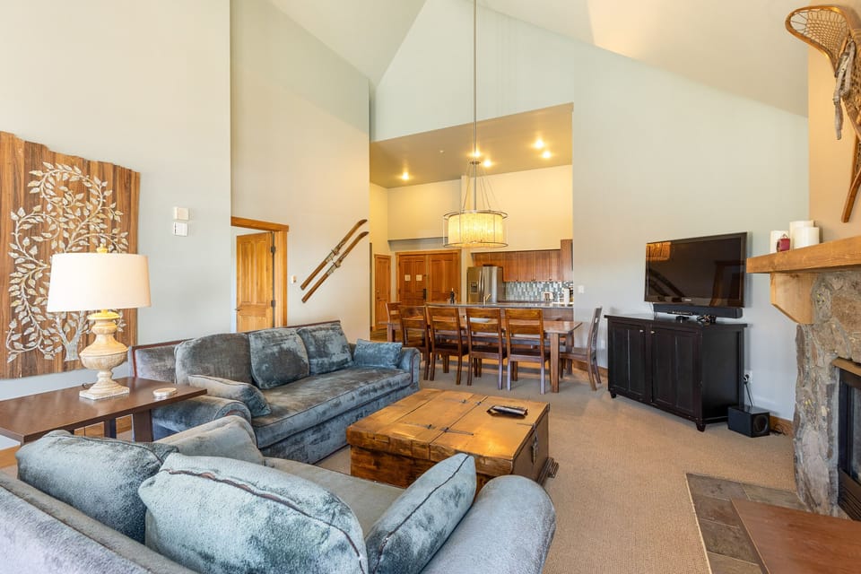 A cozy living room with a vaulted ceiling, gray couches, a wooden coffee table, a wall-mounted TV, dining table, and a stone fireplace. The room features rustic decor and a view into the kitchen area.