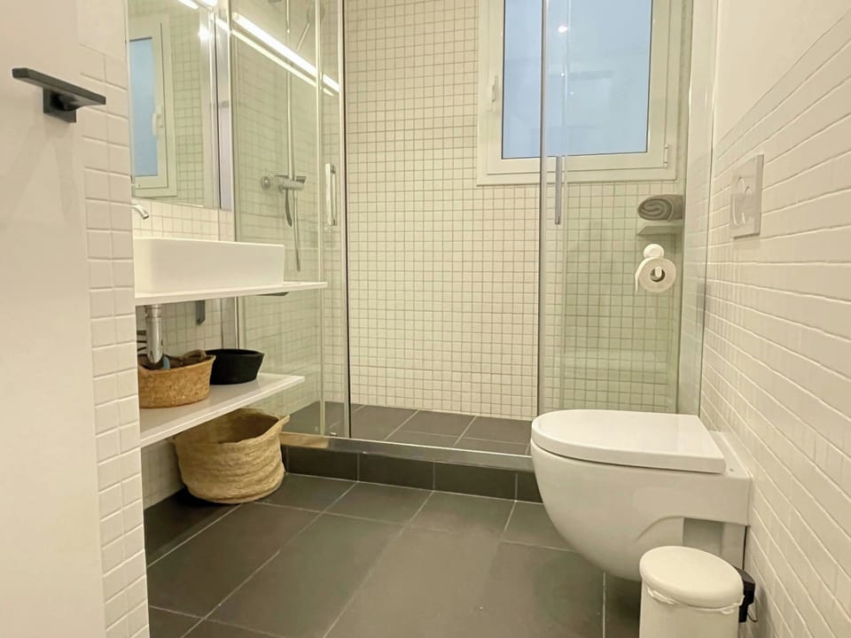 The bathroom features a modern design with white subway tiles and a sleek glass shower enclosure. The spacious shower includes a shower head and a shelf for toiletries. A white toilet and a small white trash can complement the minimalist aesthetic.  The g