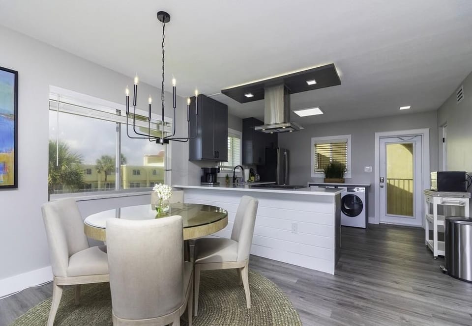 Dining area features a round glass-top table surrounded by four beige upholstered chairs on a woven rug.