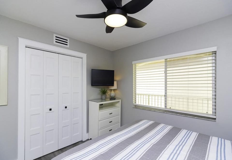 Striped gray and blue bedding covers a white bed beneath a ceiling fan in the bedroom with a window and dresser.