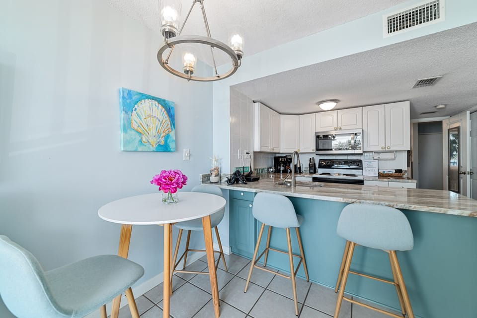 This well-appointed kitchen includes modern appliances, a large granite countertop, and a casual breakfast bar with seating. An adjacent bistro table provides additional dining space, all set within a serene, light-filled environment.
