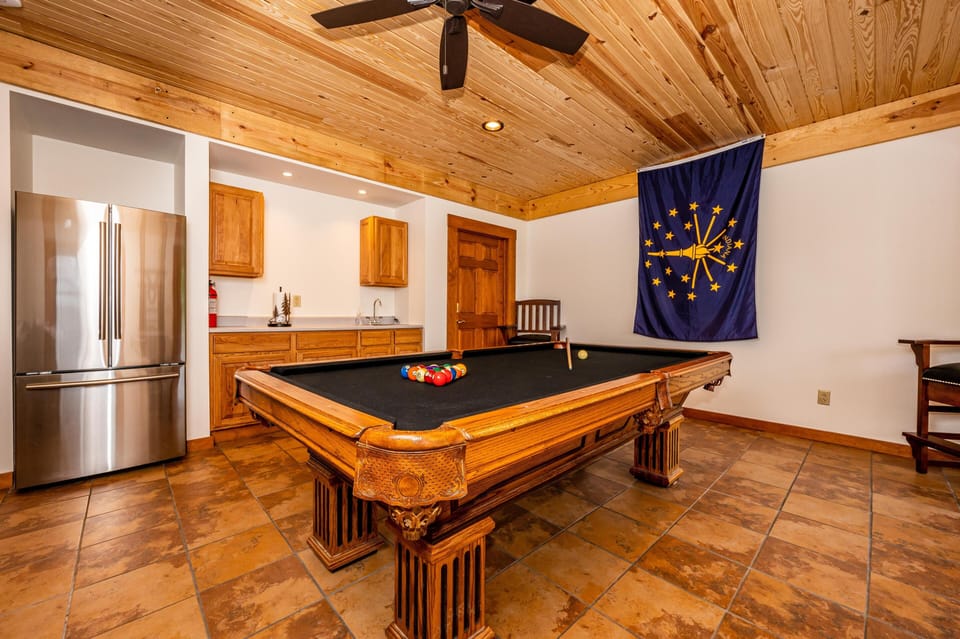 Game room with carved wood pool table, black felt, stainless steel refrigerator, and Indiana state flag on white wall.