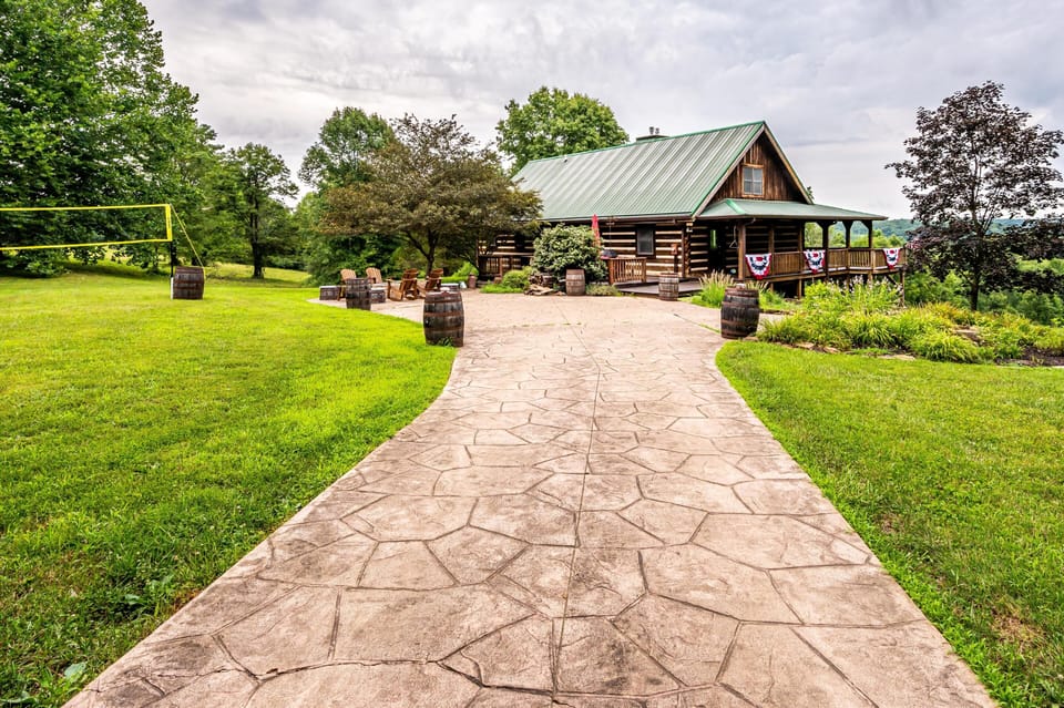 Stamped concrete driveway leads to a log cabin with green metal roof, surrounded by grass, trees, and a volleyball net.