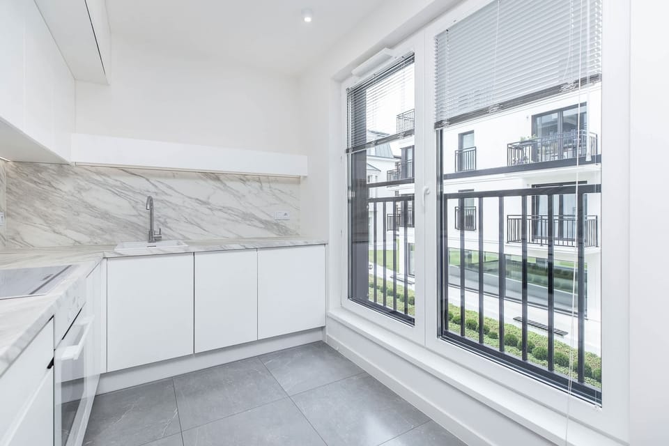 A sleek white kitchen featuring modern cabinetry, large windows with black frames, and light gray tiled flooring for a bright, contemporary cooking space.