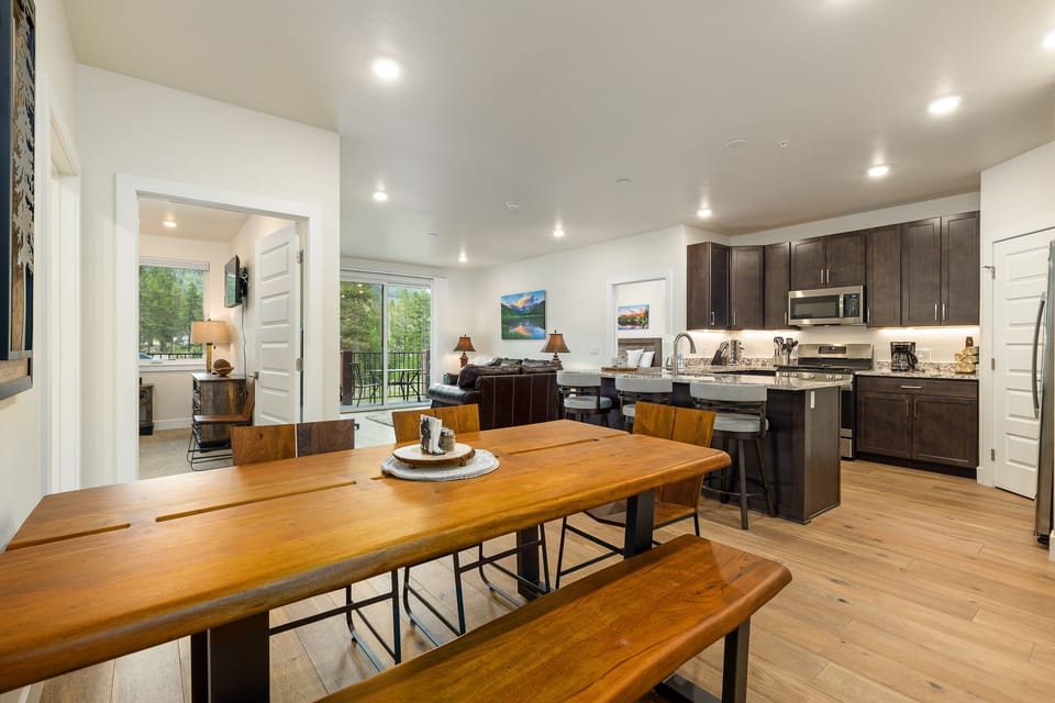 A modern kitchen and dining room with wooden furniture, stainless steel appliances, dark cabinets, and a window view of a green outdoors.