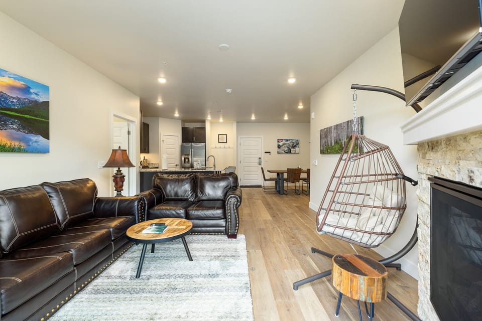 A modern living room with wooden flooring, leather sofas, a hanging chair, a coffee table, a fireplace, and a view into an open kitchen in the background.